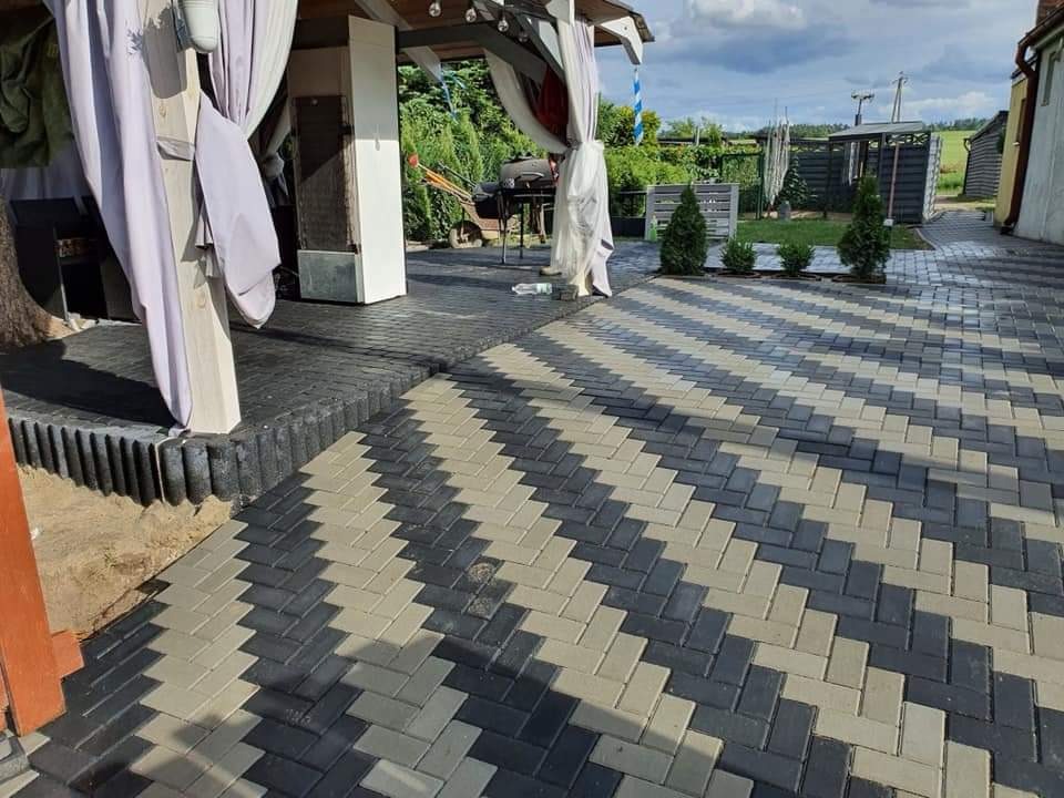 Ułożona kostka brukowa w zygzak z szarych i czarnych kostek prowadząca do zadaszonego tarasu z białymi zasłonami, w tle widoczne krzewy i budynki.