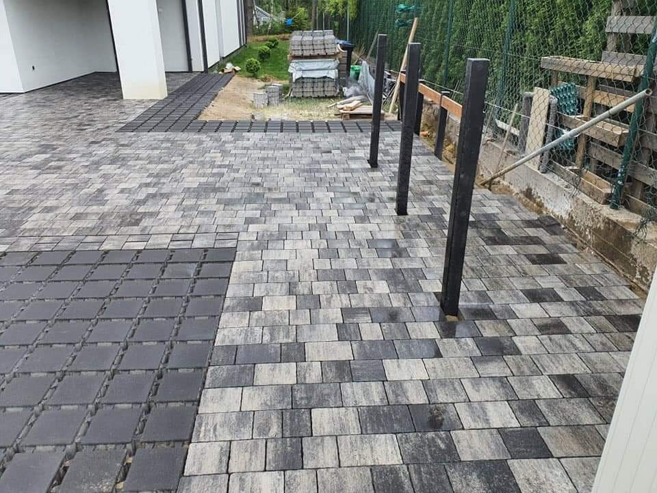 Fragment podjazdu z ułożoną szaro-grafitową kostką brukową o nieregularnym kształcie, widoczne słupki ogrodzeniowe w trakcie montażu i przygotowany teren pod dalsze prace.