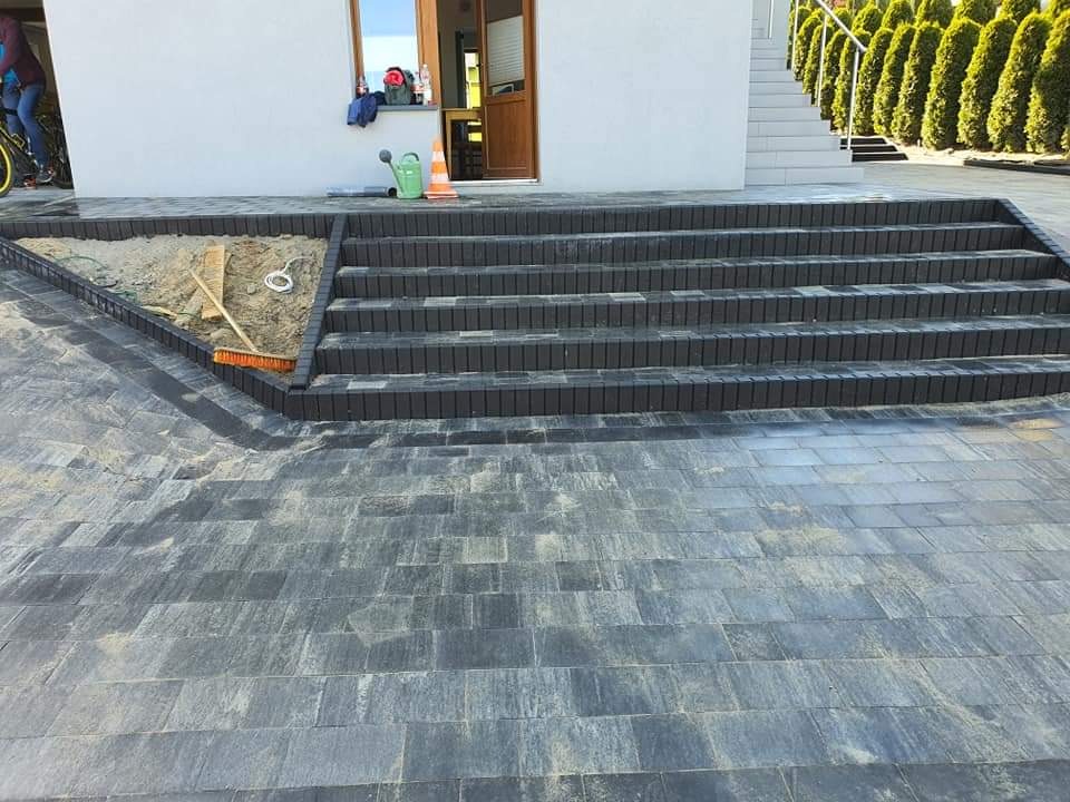 Ułożona nawierzchnia z kostki brukowej w odcieniach szarości, prowadząca do czterech stopni schodów z ciemnoszarej kostki; widoczne narzędzia brukarskie i piasek.