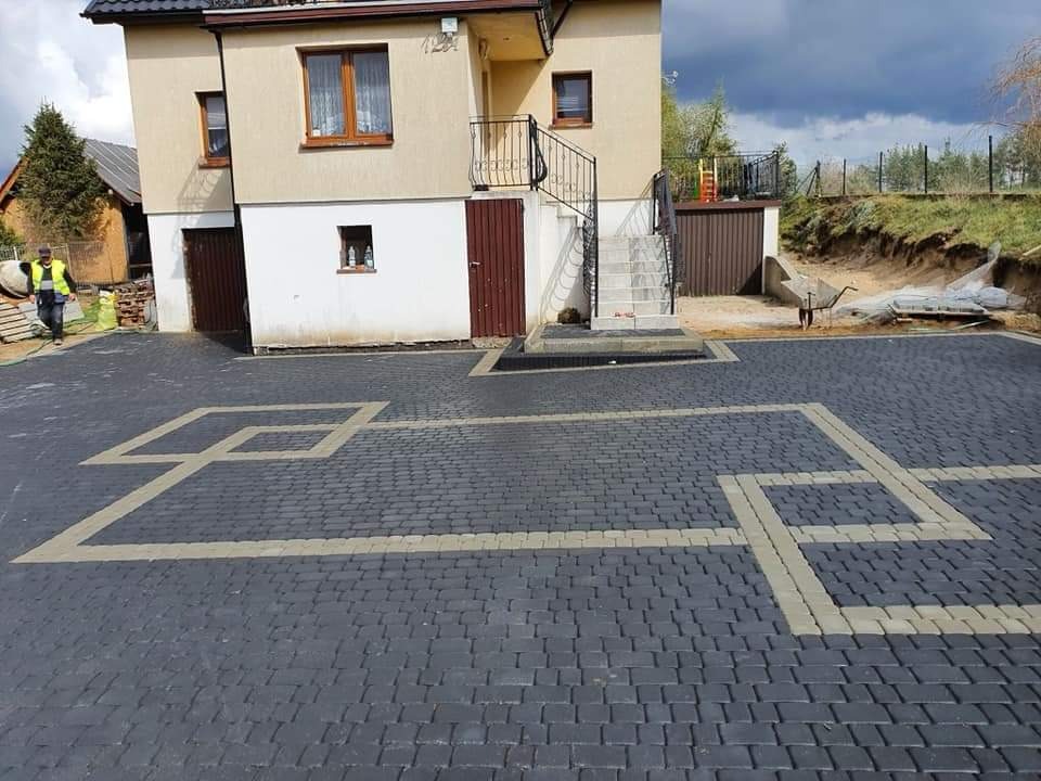 Nowa, ciemnoszara nawierzchnia z kostki brukowej ułożona przed domem, z jasnymi liniami tworzącymi geometryczny wzór. W tle widoczny pracownik w kamizelce odblaskowej, dom z zewnętrznymi schodami...