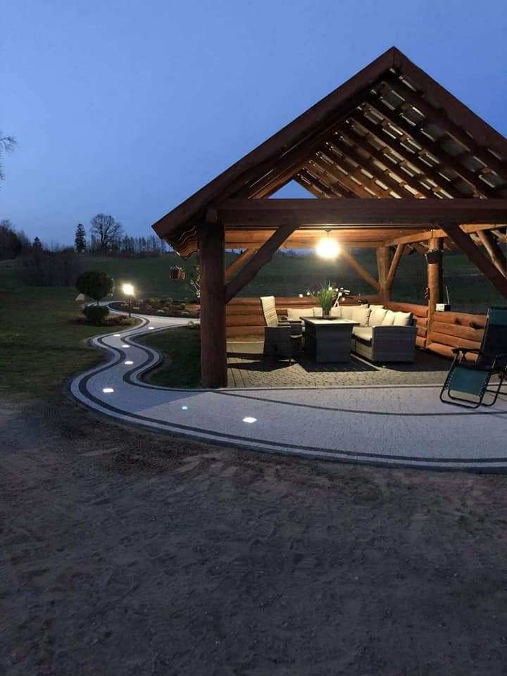 Oświetlona ścieżka z kostki brukowej prowadząca do drewnianej altany z meblami ogrodowymi, widok wczesnym wieczorem.