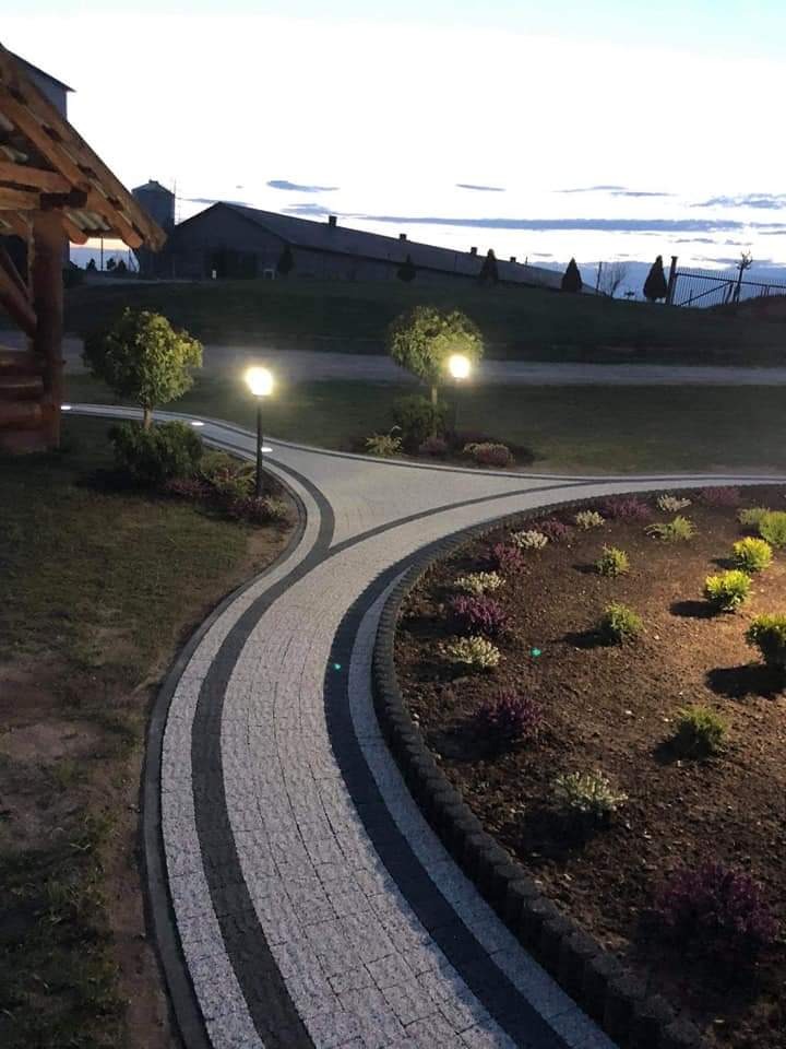 Zakrzywiony chodnik z kostki brukowej w odcieniach szarości, oświetlony niskimi lampami, prowadzący przez ogród z rabatami kwiatowymi, z widokiem na budynki w tle.