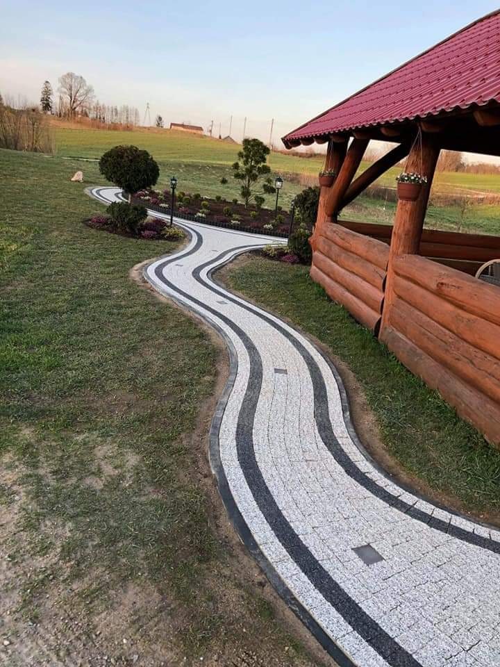 Kręta ścieżka z jasnej kostki brukowej z czarnymi pasami, prowadząca przez trawnik w kierunku altany z czerwonym dachem, oświetlona lampami ogrodowymi.