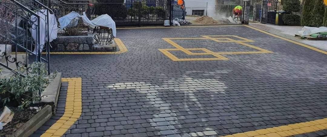 Ułożona, szara kostka brukowa z żółtymi akcentami w kształcie kwadratów, widoczni pracownicy w tle, brama wjazdowa.