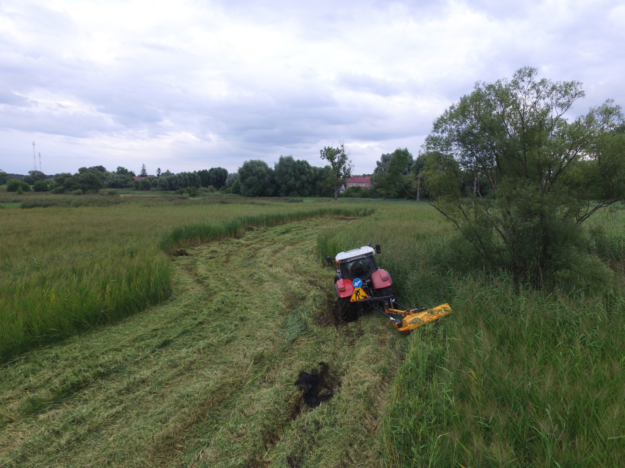 Czerwony traktor z żółtą kosiarką rotacyjną tnie pas trawy na zarośniętym polu, widoczne drzewa i budynki w tle pod pochmurnym niebem.