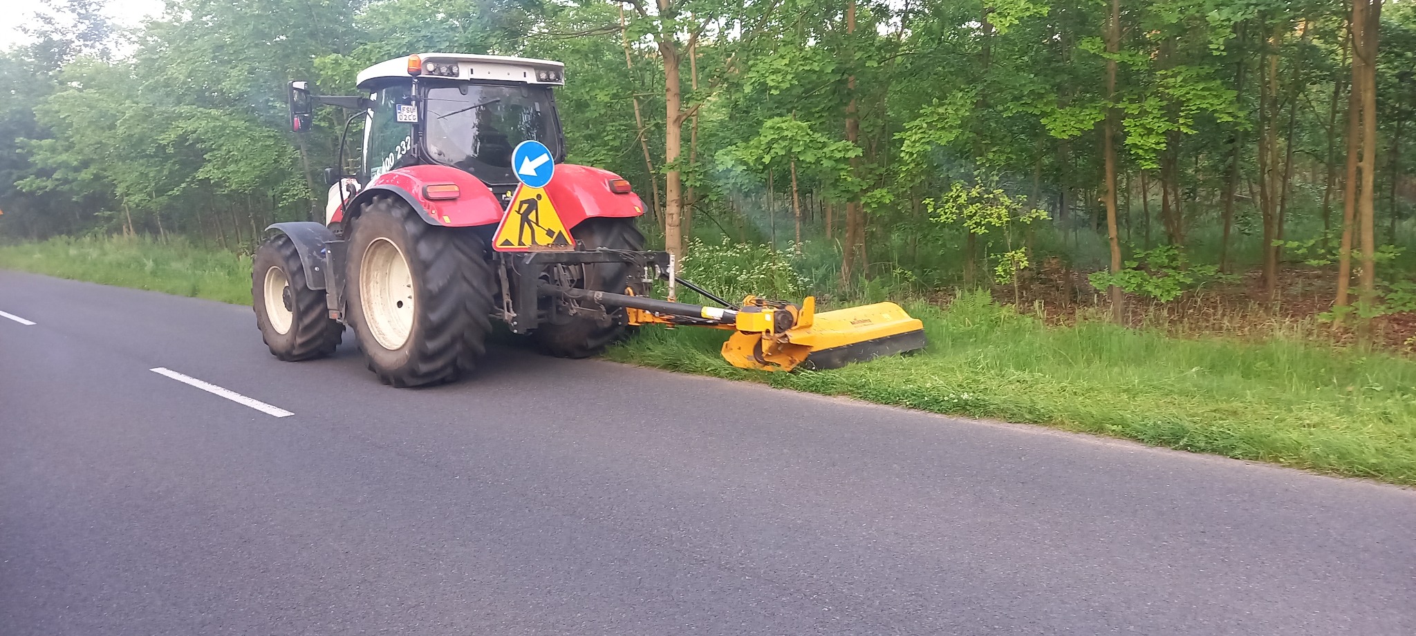 Czerwony traktor z żółtą kosiarką boczną przycinający trawę na poboczu asfaltowej drogi, w tle drzewa i znak ostrzegawczy o robotach drogowych.