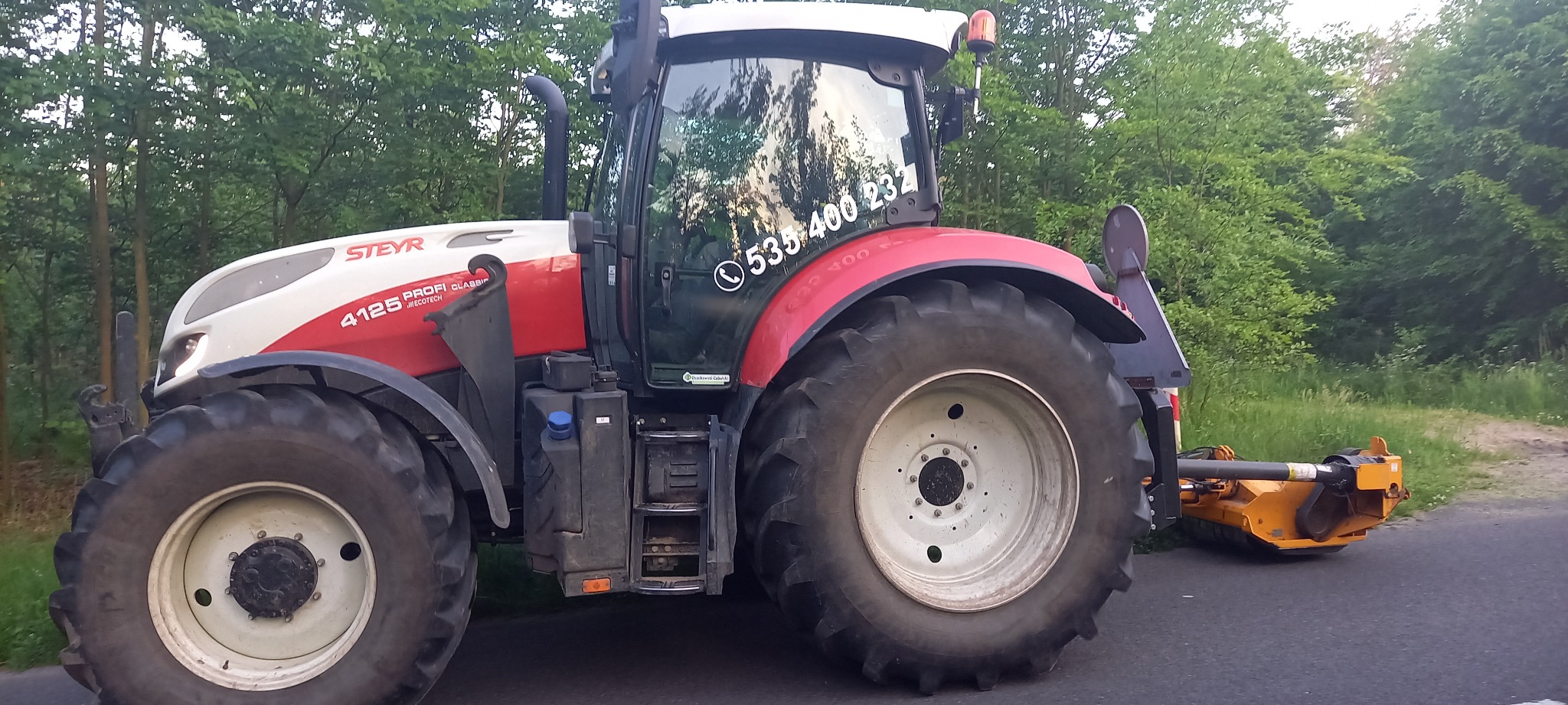 Czerwono-biały traktor Steyr 4125 Profi Classic z numerem telefonu na szybie, zaparkowany na asfaltowej drodze obok zarośli i lasu, z podłączoną żółtą kosiarką bijakową.