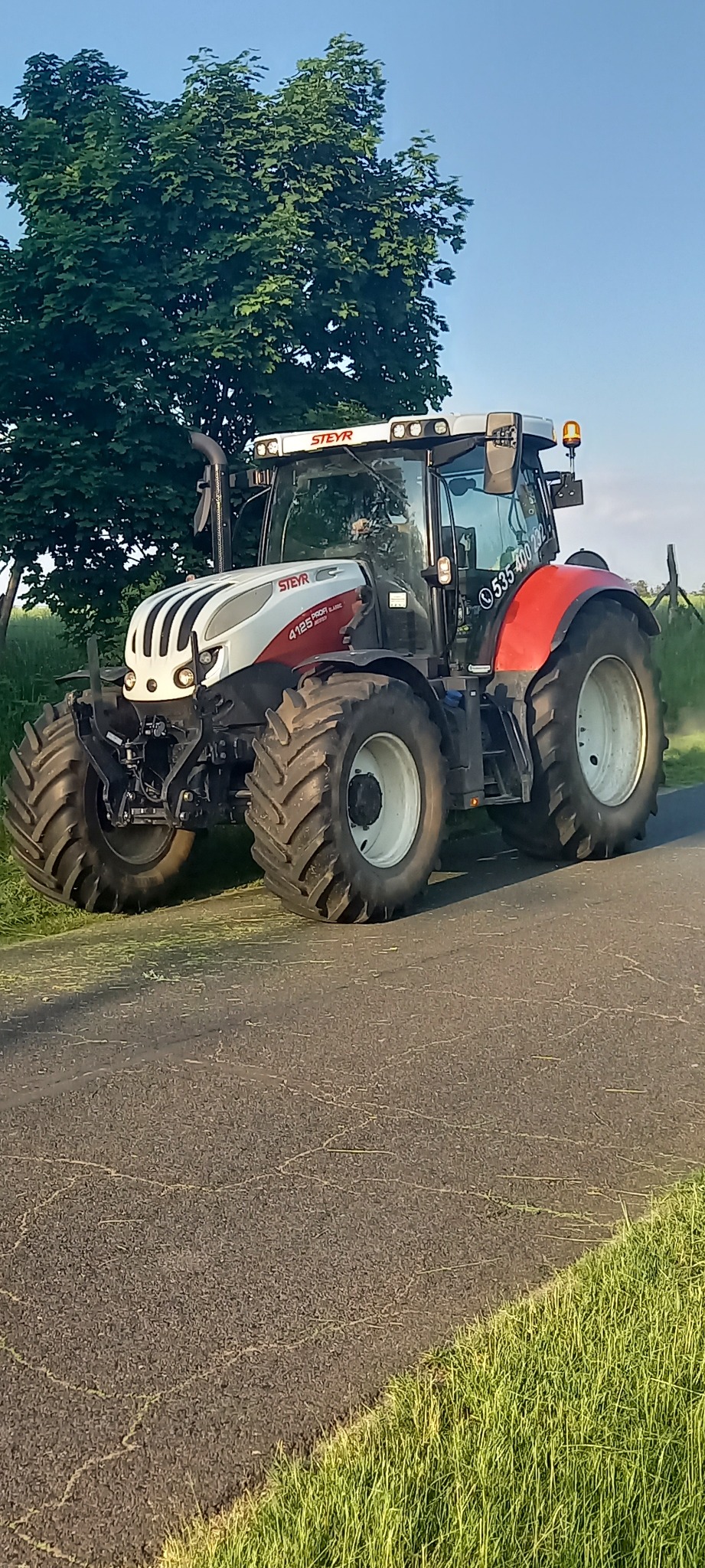 Czerwono-biały traktor Steyr 4125 Profi Classic na asfaltowej drodze, z zielonym drzewem w tle i numerem telefonu na kabinie.
