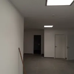 Wnętrze korytarza z szarą podłogą z płytek, białe ściany, sufit podwieszany z oświetleniem LED i widoczną drewnianą listwą przypodłogową opartą o ścianę.