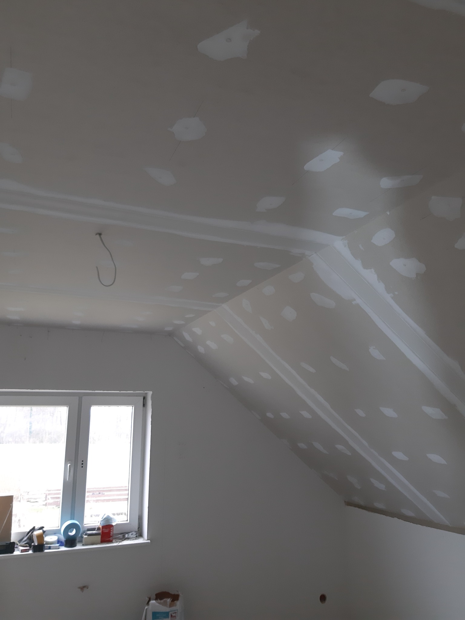 Poddasze po montażu płyt kartonowo-gipsowych, widoczne szpachlowane łączenia i wkręty, okno z narzędziami na parapecie, surowy kabel zwisający z sufitu.