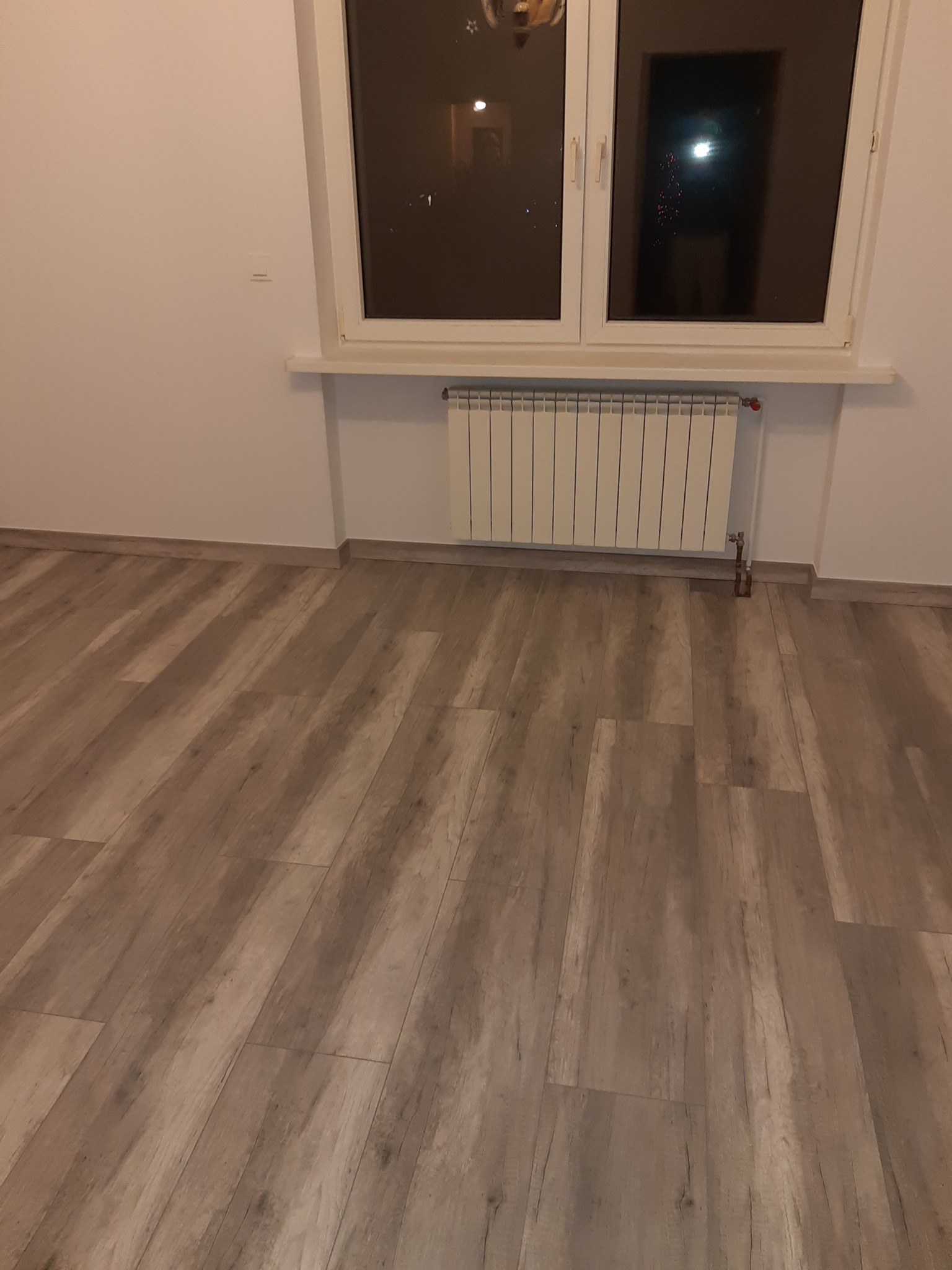 Pusty pokój po remoncie z panelami podłogowymi w odcieniach brązu, białymi ścianami i grzejnikiem pod oknem z widokiem na nocne miasto.