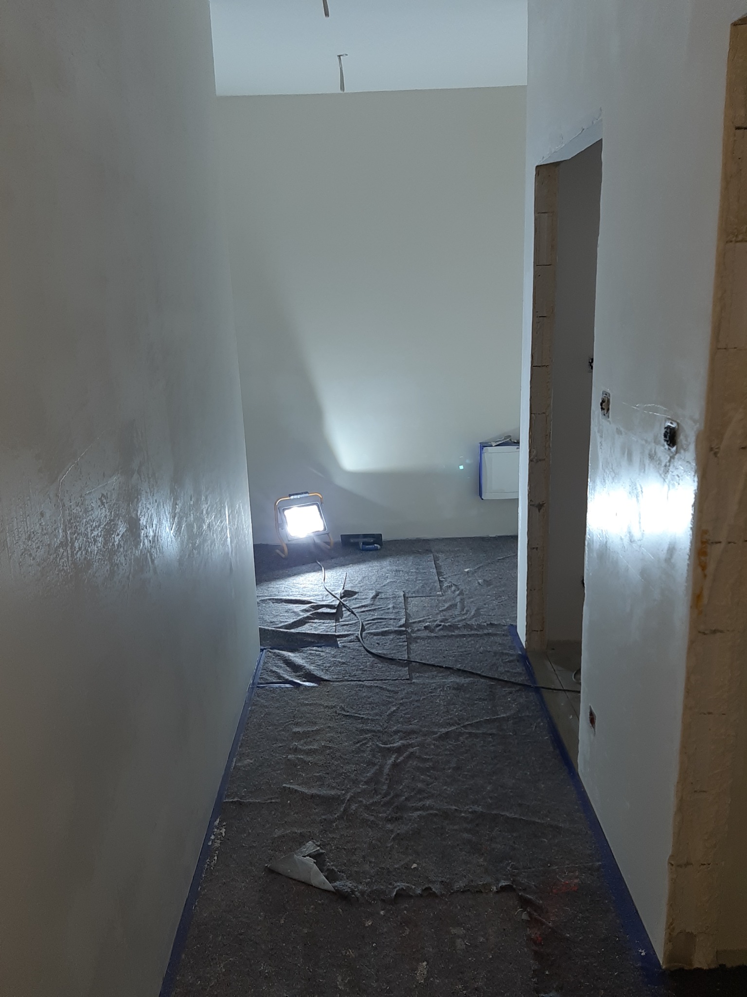 Pokój w trakcie prac wykończeniowych, zabezpieczona podłoga, świeżo pomalowane ściany oświetlone lampą budowlaną, widoczne przygotowane otwory na gniazdka.