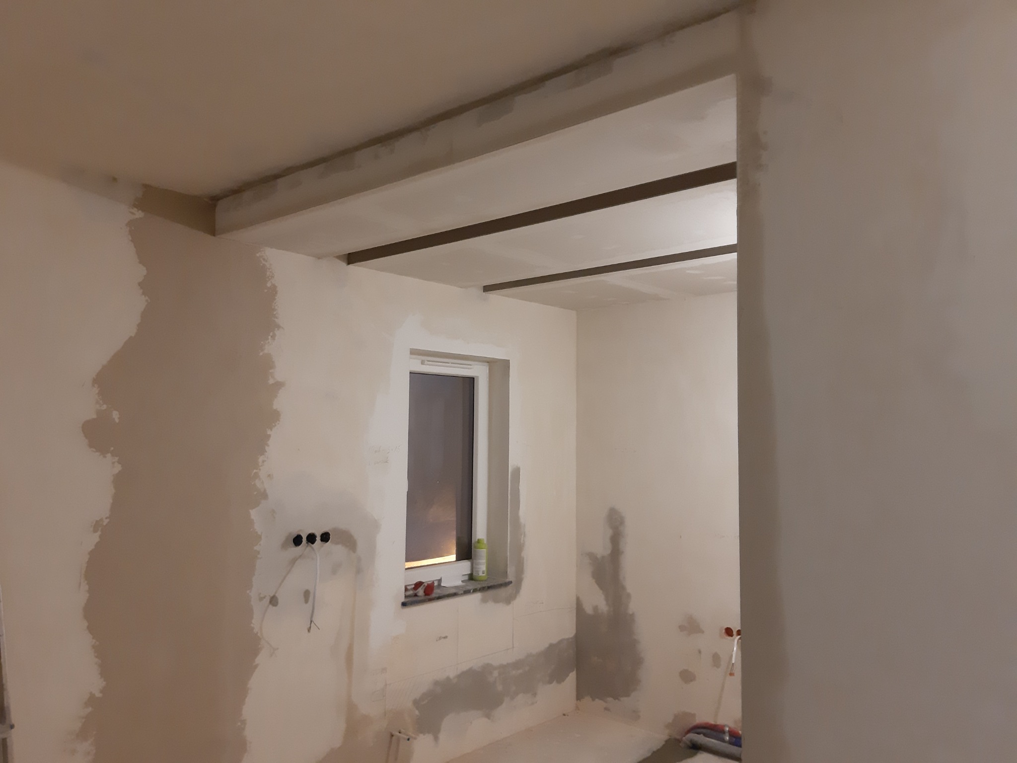 Wnętrze w trakcie prac wykończeniowych: surowe ściany z widocznymi śladami szpachlowania, przygotowane pod malowanie, widoczne kable elektryczne, okno z parapetem, konstrukcja sufitu podwieszanego...