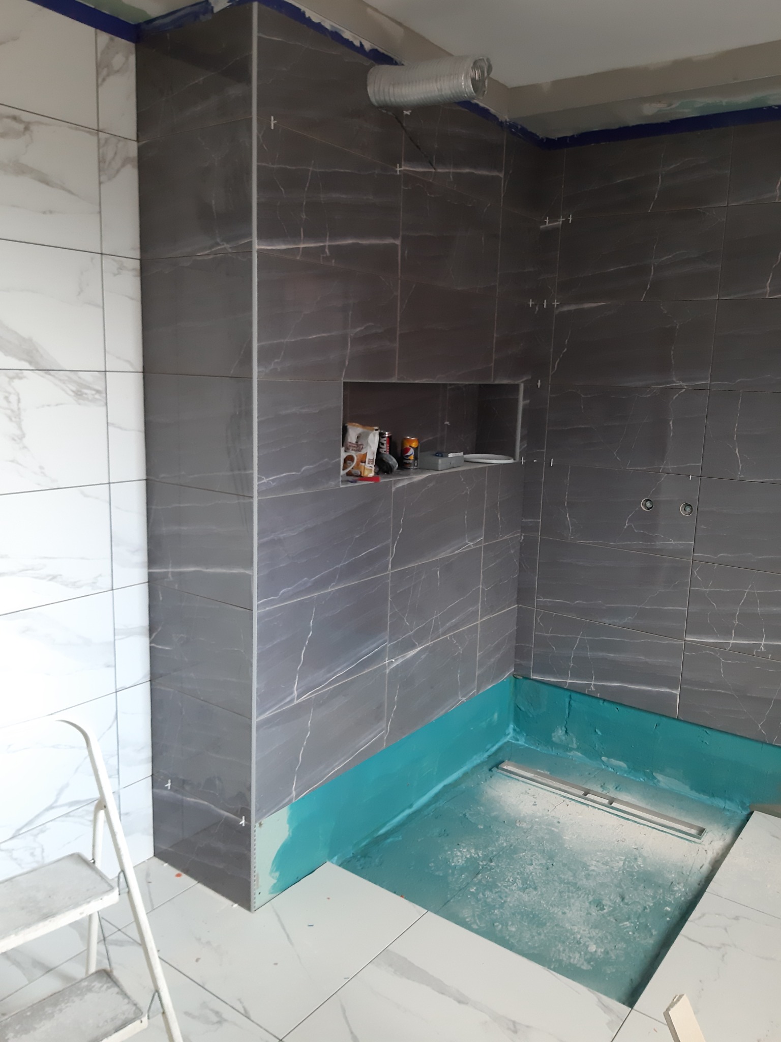 Montaż płytek w łazience: szare płytki imitujące marmur na ścianach, białe płytki na podłodze, widoczny brodzik z odpływem liniowym i hydroizolacją w kolorze turkusowym, drabinka malarska w rogu.