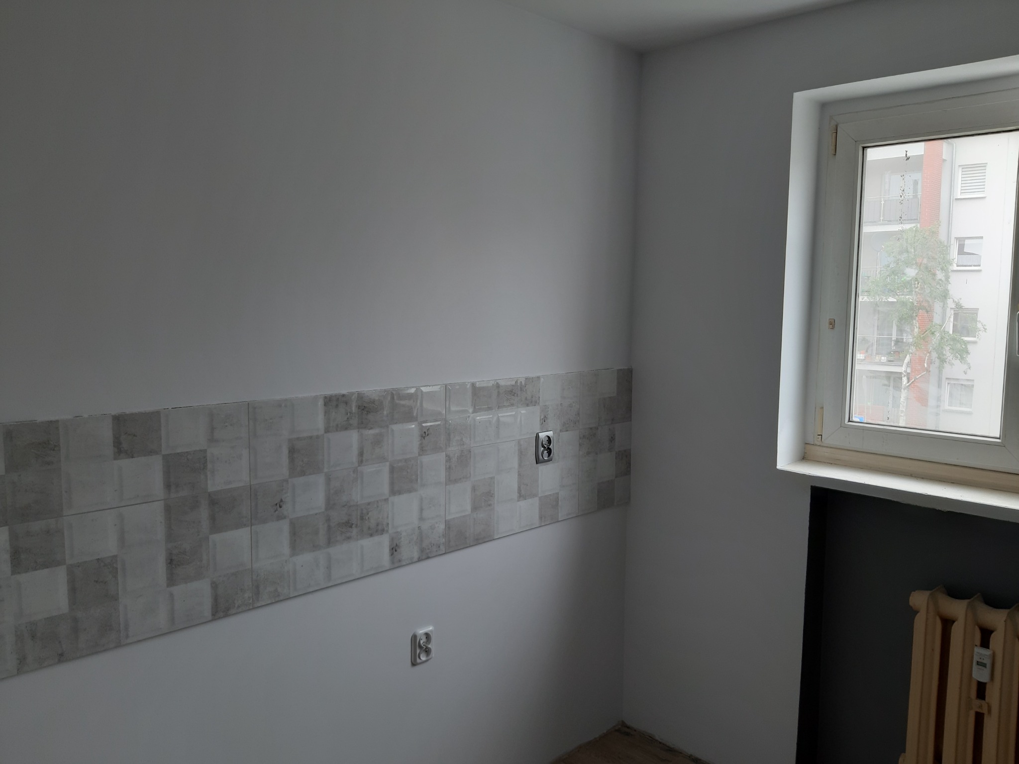 Fragment ściany z ułożoną szarą mozaiką kwadratową, gniazdka elektryczne, okno z widokiem na budynek i drzewo, oraz grzejnik żeberkowy w rogu pomieszczenia.