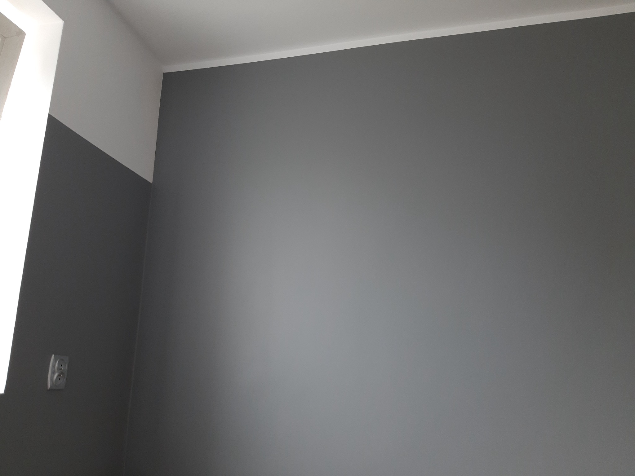 Szara ściana pomalowana do pewnej wysokości, a powyżej biała, z widocznym gniazdkiem elektrycznym i listwą przysufitową.