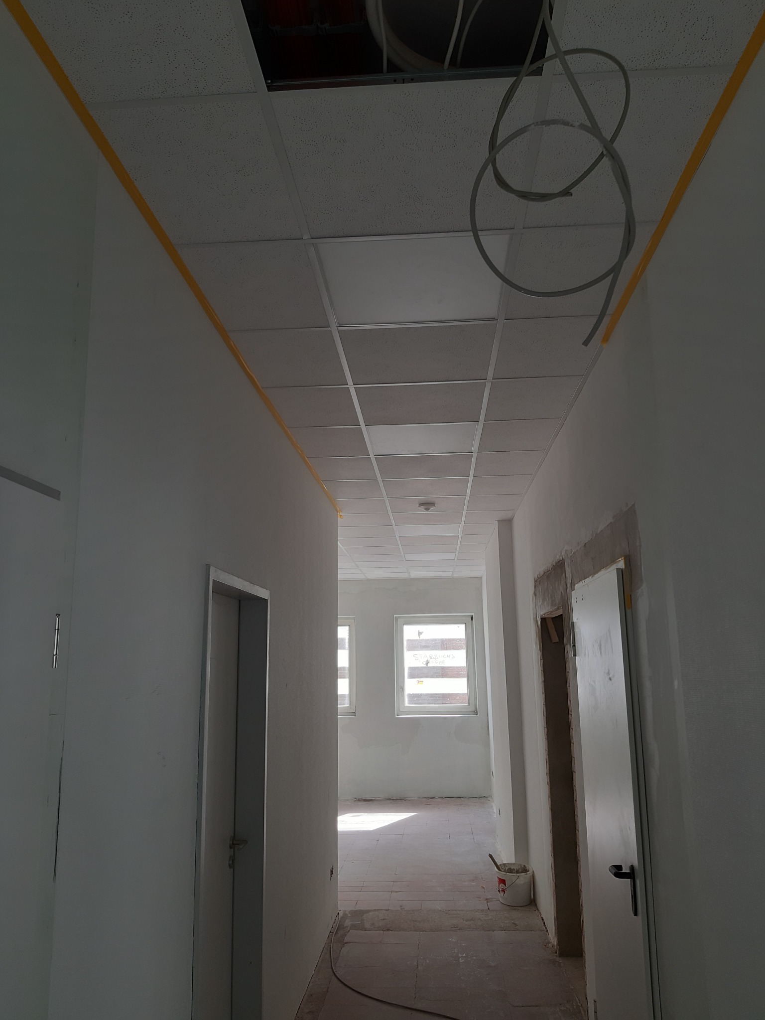 Korytarz w trakcie prac wykończeniowych, widoczne ślady po taśmie malarskiej na ścianach, podwieszany sufit z otwartym fragmentem, wystające przewody oraz wiadro z narzędziami na podłodze.
