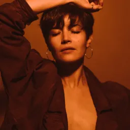 Portret kobiety z krótkimi włosami, z zamkniętymi oczami, w brązowej kurtce, na jednolitym, ciepłym tle, z kolczykami kołami i pierścionkiem na palcu.