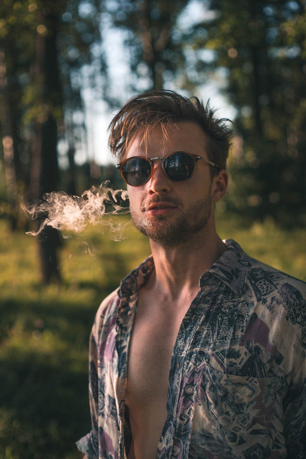 Młody mężczyzna w okularach przeciwsłonecznych, z rozpiętą koszulą w etniczne wzory, wypuszcza dym na tle zieleni.