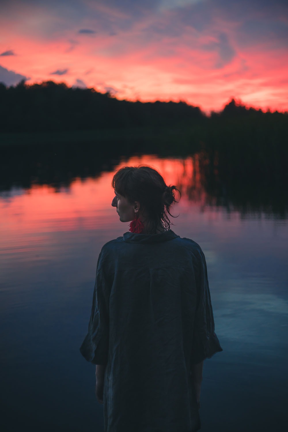 Kobieta z upiętymi włosami i czerwonym kolczykiem, stojąca tyłem nad jeziorem o zmierzchu z odbijającym się niebem w wodzie.