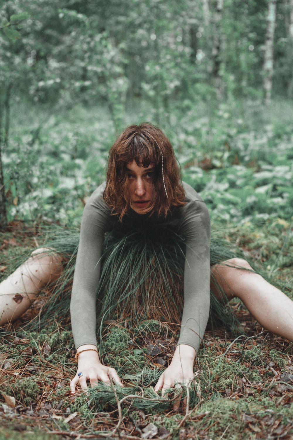 Kobieta w zielonym stroju, siedząca w lesie, otoczona trawą i mchem, z wyrazistym spojrzeniem skierowanym w obiektyw.