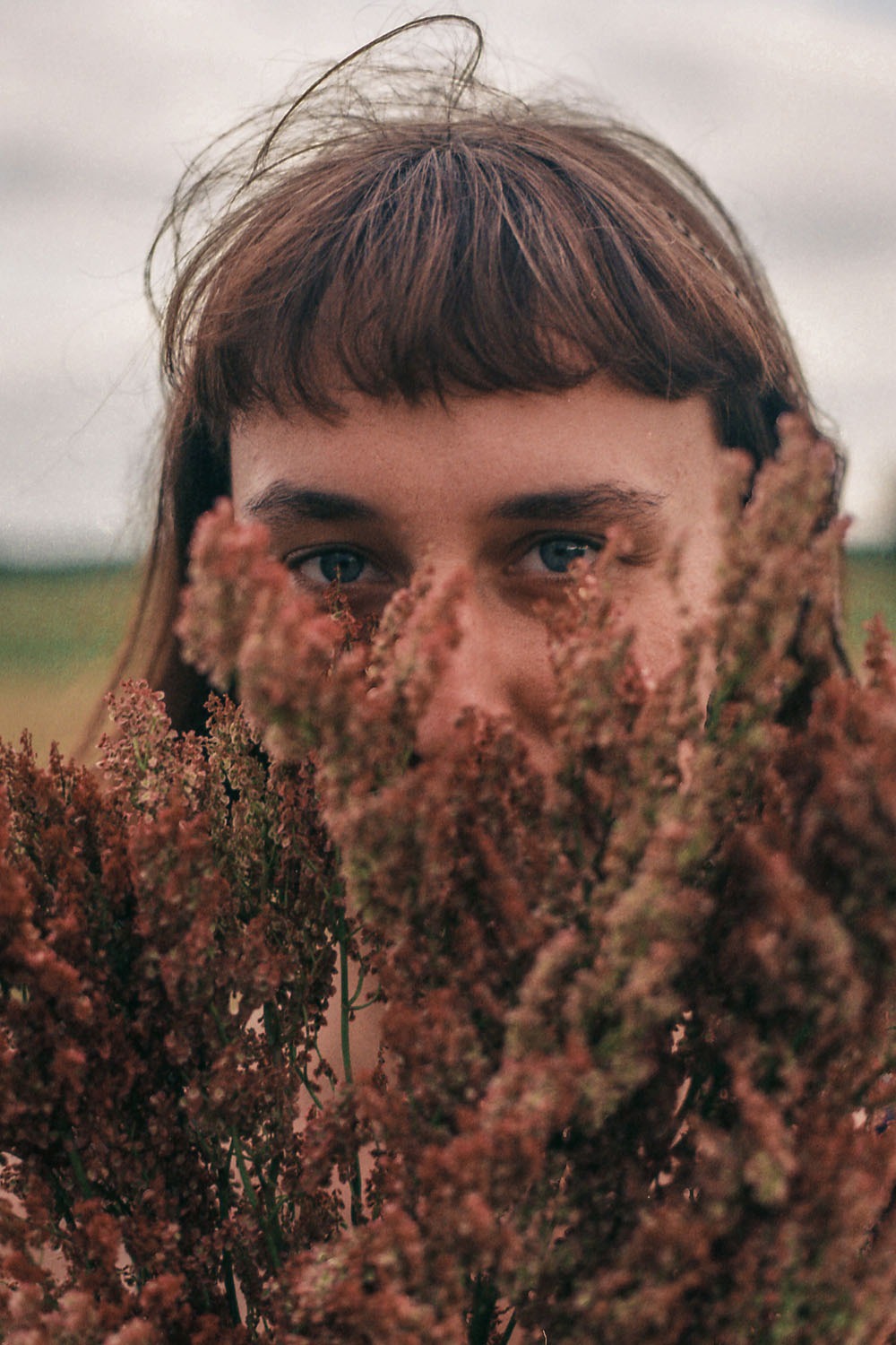 Portret kobiety o jasnych oczach, częściowo zasłoniętej przez bukiet brązowo-czerwonych kwiatów polnych, z rozwichrzonymi włosami na tle rozmytego pola.