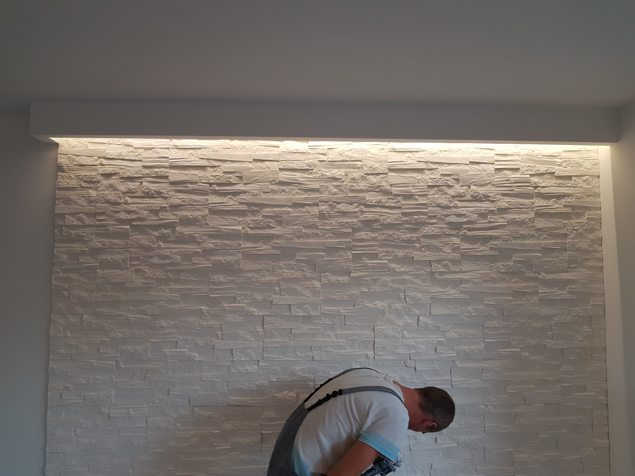 Pracownik w kombinezonie montuje dekoracyjną, białą ścianę z paneli imitujących kamień, oświetloną od góry listwą LED.