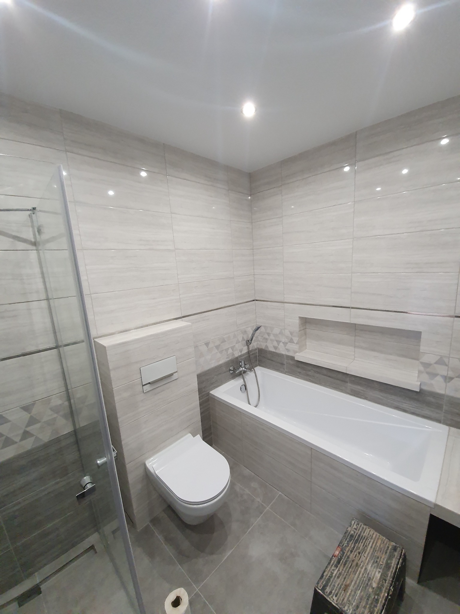 Wykończona łazienka z szarymi płytkami imitującymi drewno, kabina prysznicowa ze szkła hartowanego, podwieszana toaleta i wanna z zabudową, widoczny stołek roboczy.