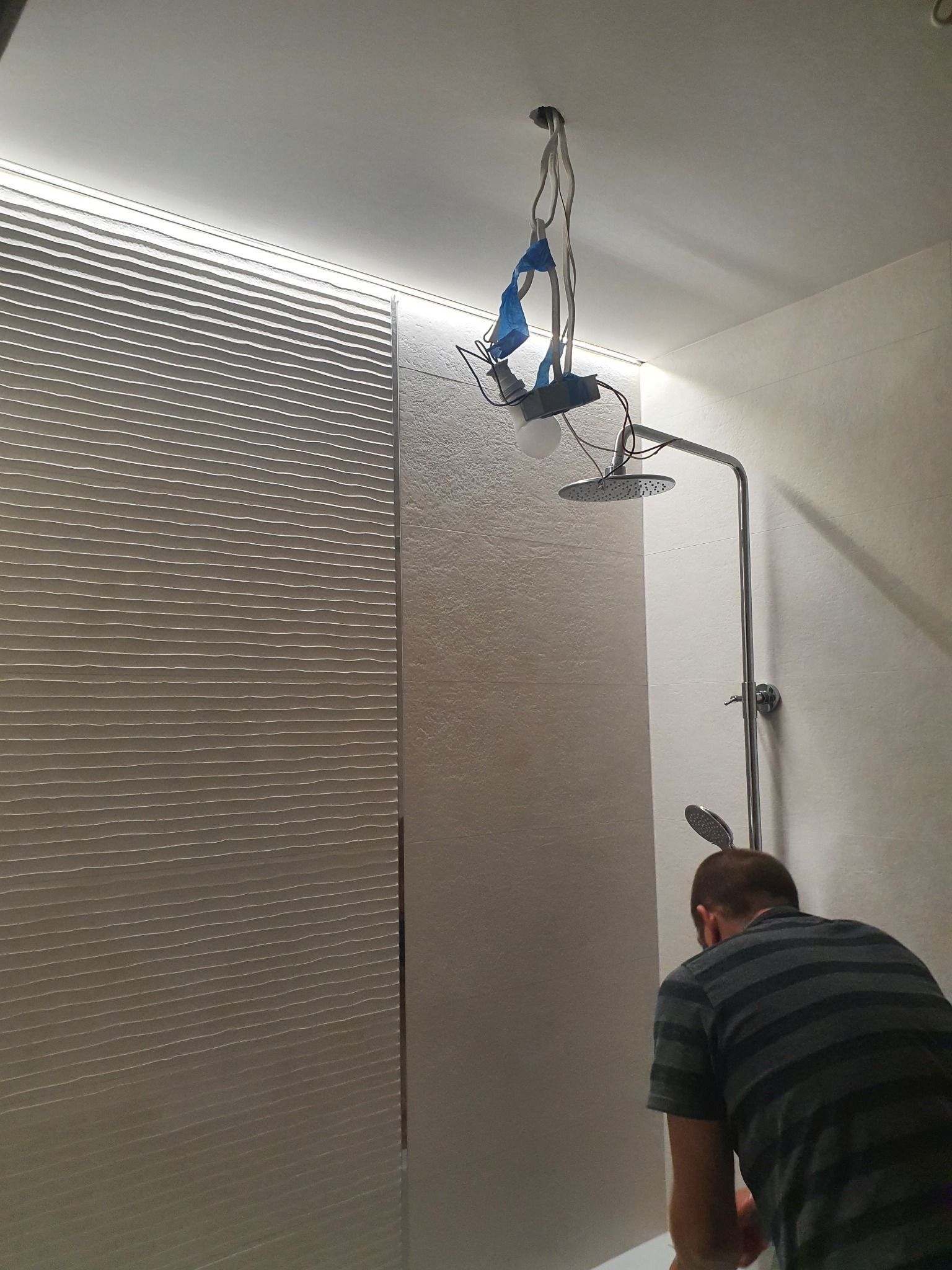 Instalacja deszczownicy w łazience z podświetleniem LED; widoczne wiszące przewody i tymczasowe oświetlenie.