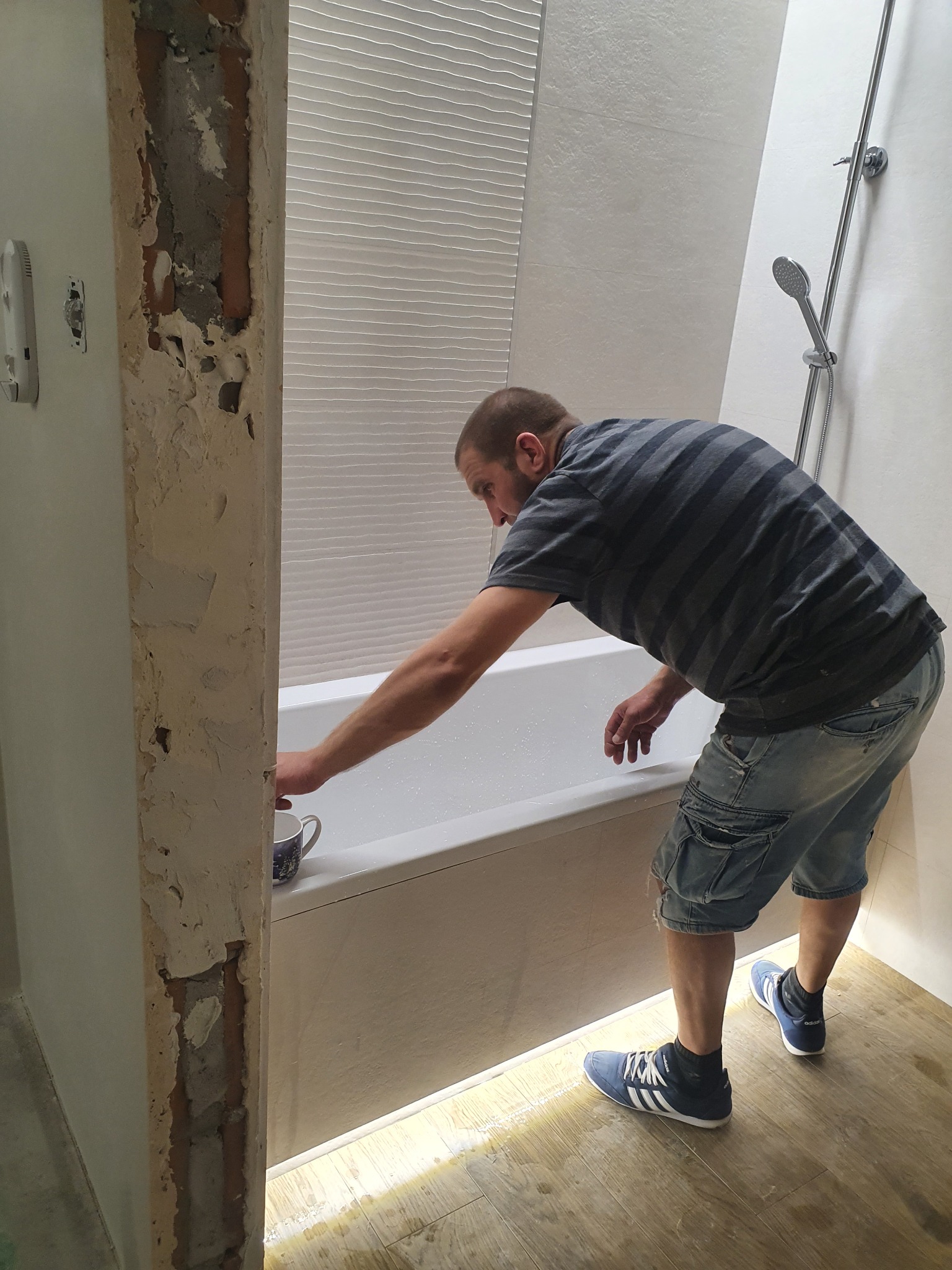 Mężczyzna w paski pochyla się nad białą wanną w trakcie remontu łazienki, widoczny fragment ściany z odsłoniętą cegłą i kubek na skraju wanny.