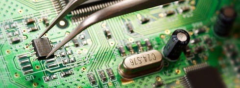 Precyzyjny montaż układu scalonego na zielonej płycie głównej za pomocą metalowej pęsety; widoczne liczne komponenty elektroniczne i ścieżki.