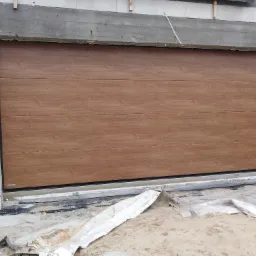 Segmentowa brama garażowa z paneli imitujących drewno, zamontowana w surowym otworze garażowym z betonu komórkowego, widoczne ślady montażu i zabezpieczenia podłoża.