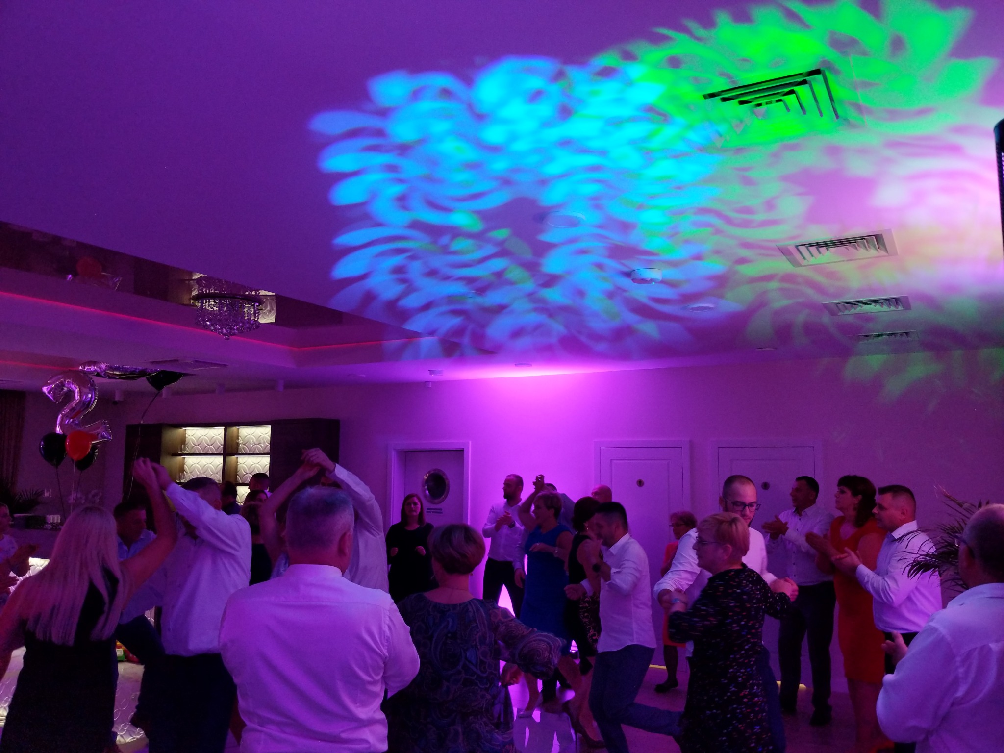 Tańczący ludzie w sali oświetlonej fioletowym światłem z dekoracyjnymi projekcjami na suficie i balonami w tle.