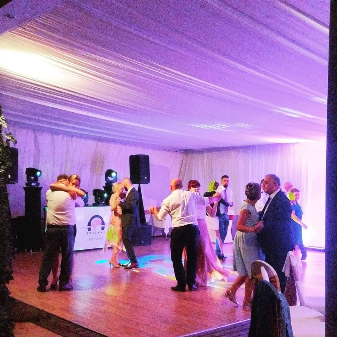 Tańczący goście weselni na parkiecie oświetlonym fioletowymi światłami, widoczne elementy nagłośnienia i dekoracje sufitowe z białego materiału.