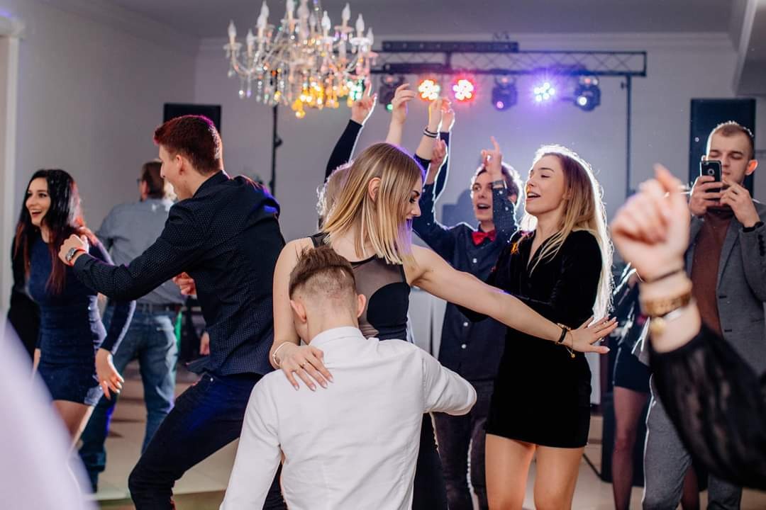 Grupa młodych osób tańczy na parkiecie podczas imprezy, oświetlonym kolorowymi światłami i pod kryształowym żyrandolem.