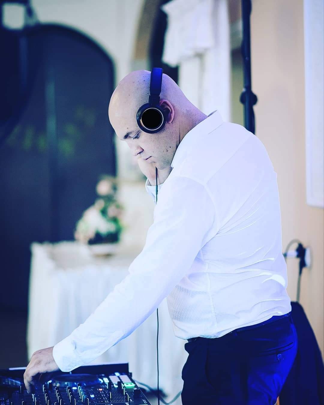 Łysy mężczyzna w białej koszuli i słuchawkach obsługuje konsoletę DJ-ską, w tle stół z dekoracją kwiatową.