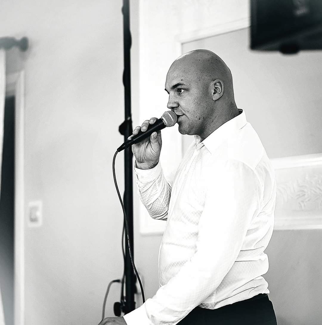 Czarno-białe zdjęcie mężczyzny w białej koszuli, trzymającego mikrofon i gotowego do śpiewania, z widocznym statywem mikrofonowym.