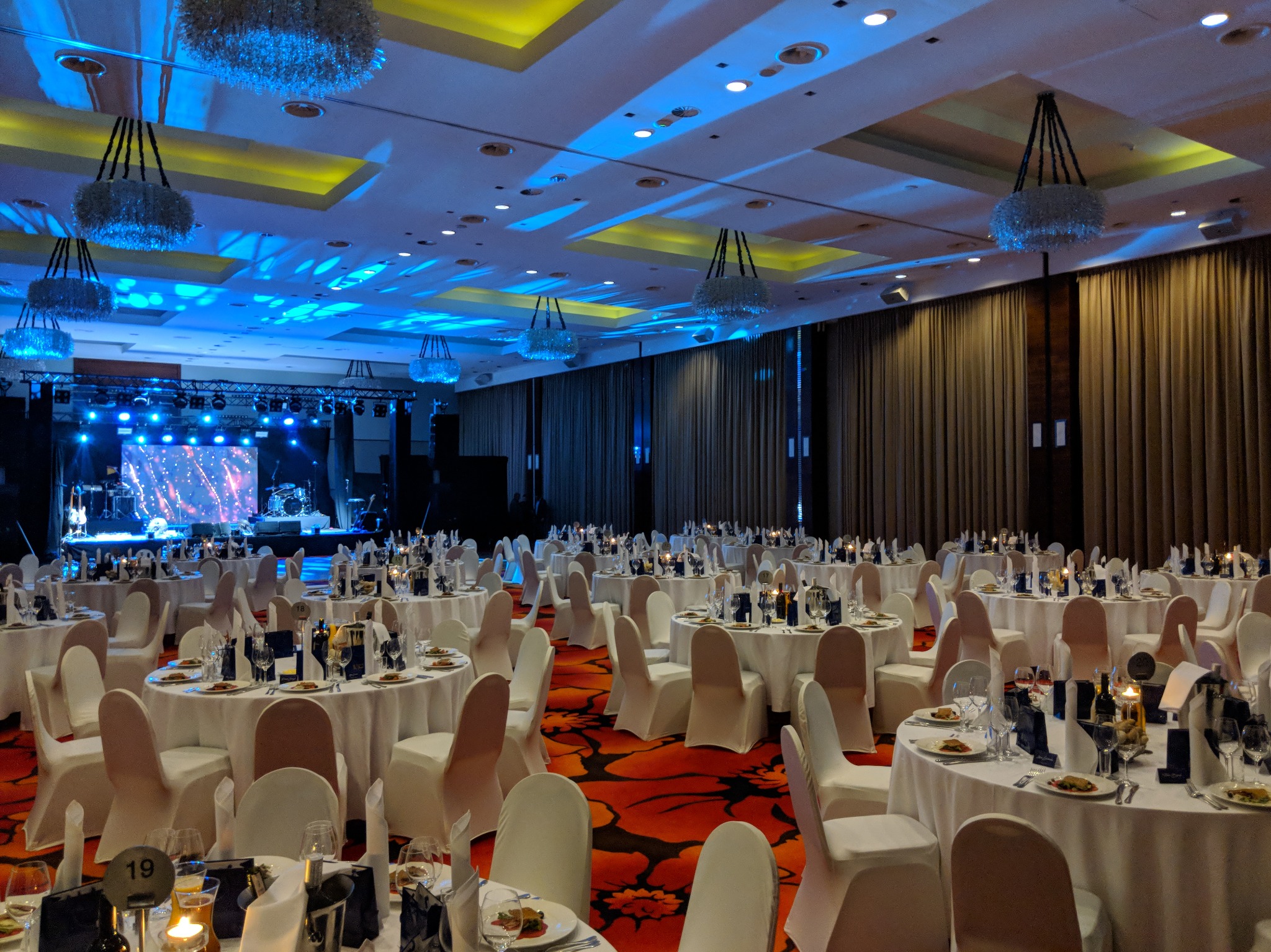 Sala bankietowa z okrągłymi stołami nakrytymi białymi obrusami, krzesłami w pokrowcach, dekoracyjnymi żyrandolami i sceną z oświetleniem w tle, przygotowana na uroczystość.