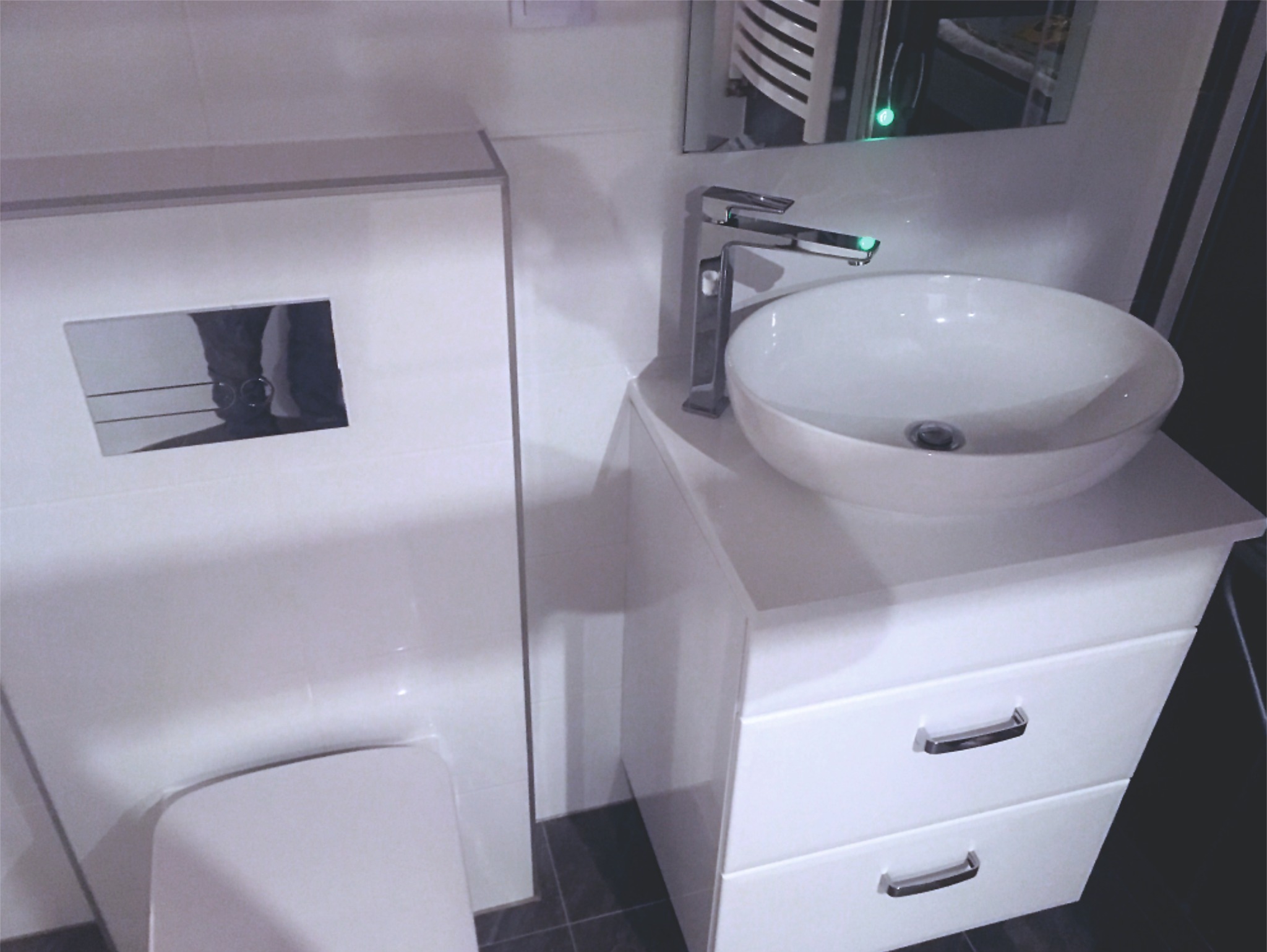 Nowoczesna, biała umywalka nablatowa z chromowaną baterią, zamontowana na szafce z dwiema szufladami, obok zabudowy WC z przyciskiem spłukującym, w łazience wykończonej białymi płytkami.