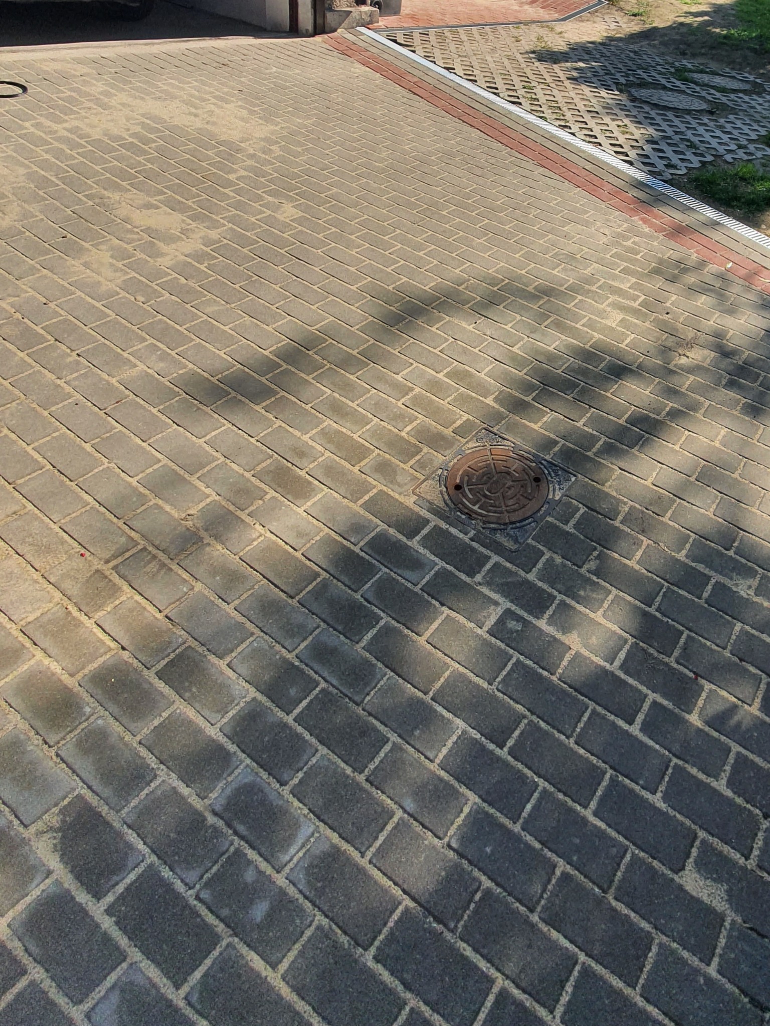 Szara kostka brukowa ułożona w jodełkę z okrągłą studzienką kanalizacyjną pośrodku, cień drzewa pada na powierzchnię.