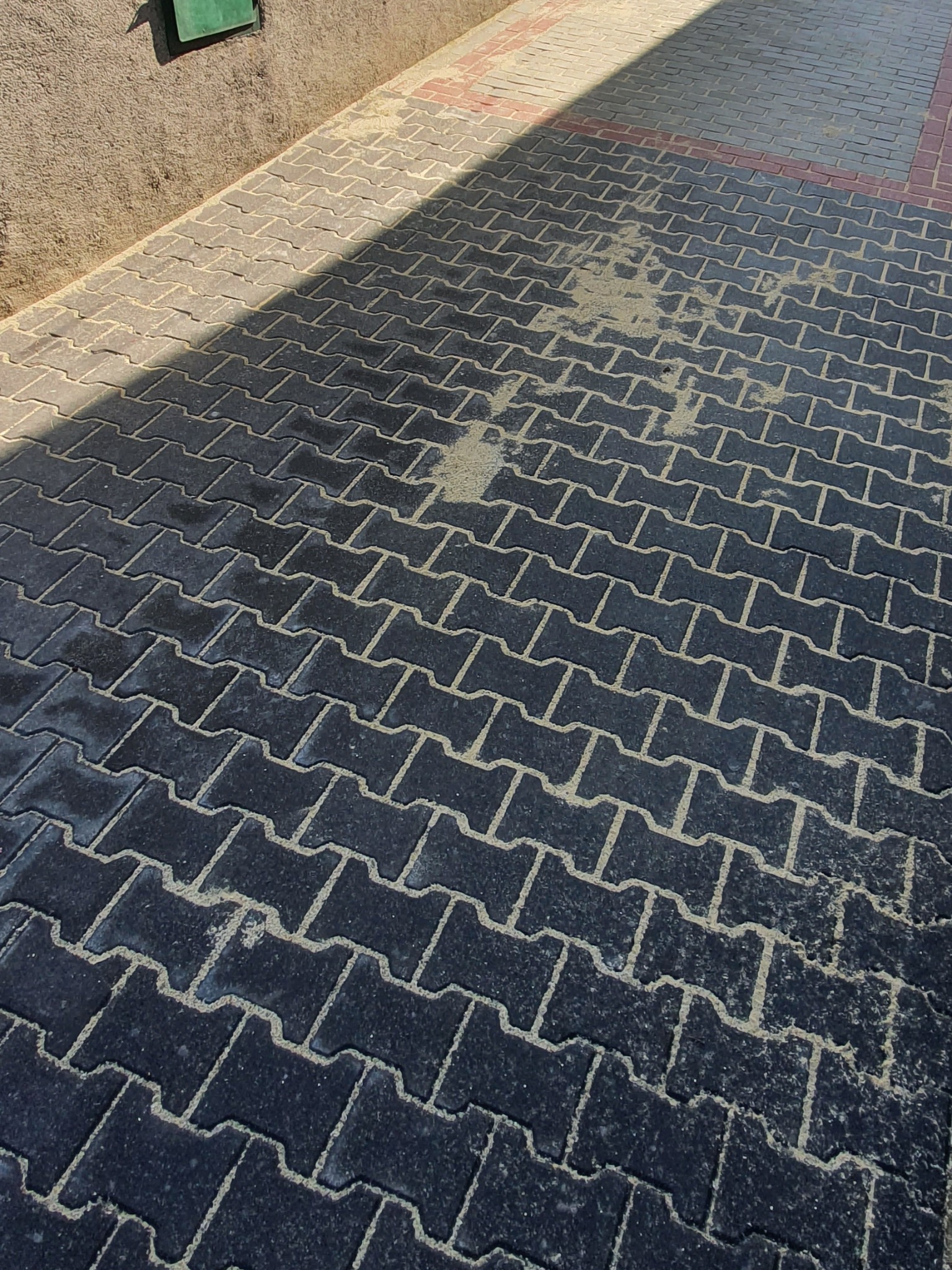 Ciemnoszara kostka brukowa ułożona w zygzak, z widocznymi śladami piasku na powierzchni i w fugach, częściowo oświetlona słońcem, przylegająca do betonowej ściany i czerwonej obwódki.