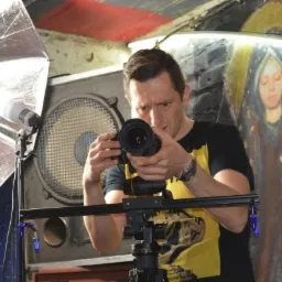 Mężczyzna z aparatem na sliderze, ustawia ostrość, w tle lampa błyskowa i malowidło na ścianie.