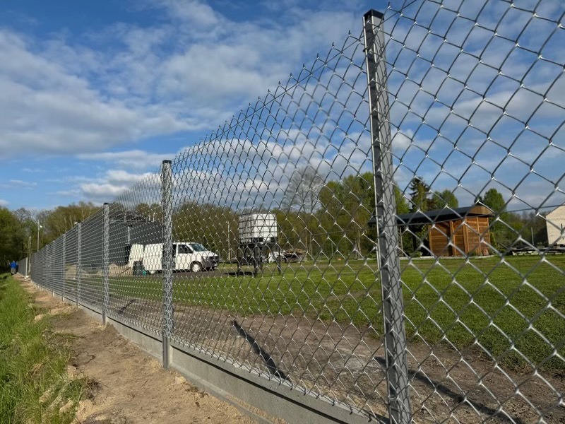 Długi odcinek ogrodzenia z siatki na betonowej podmurówce, widok z zewnątrz posesji, z fragmentem trawnika i zabudowaniami w tle.