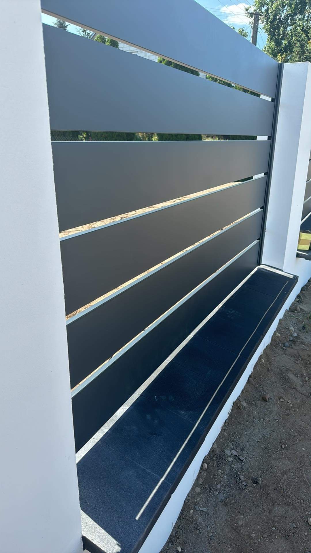 Nowoczesne poziome przęsła w kolorze grafitowym osadzone na białych słupkach, widok z bliska na fragment ogrodzenia z czarną podmurówką.