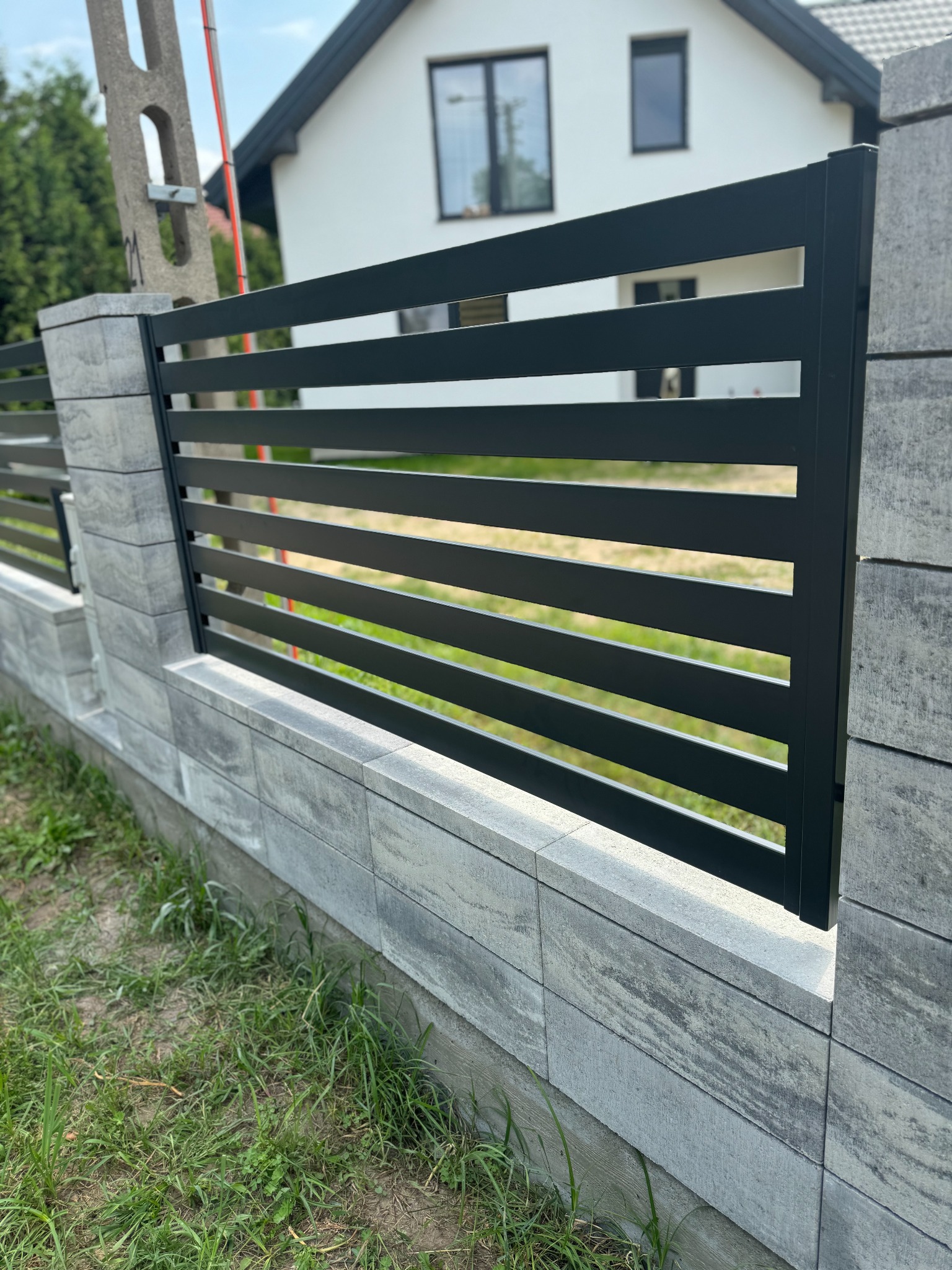 Nowoczesne ogrodzenie z poziomych, czarnych profili zamontowane na szarym, betonowym murku z widocznymi elementami domu w tle.