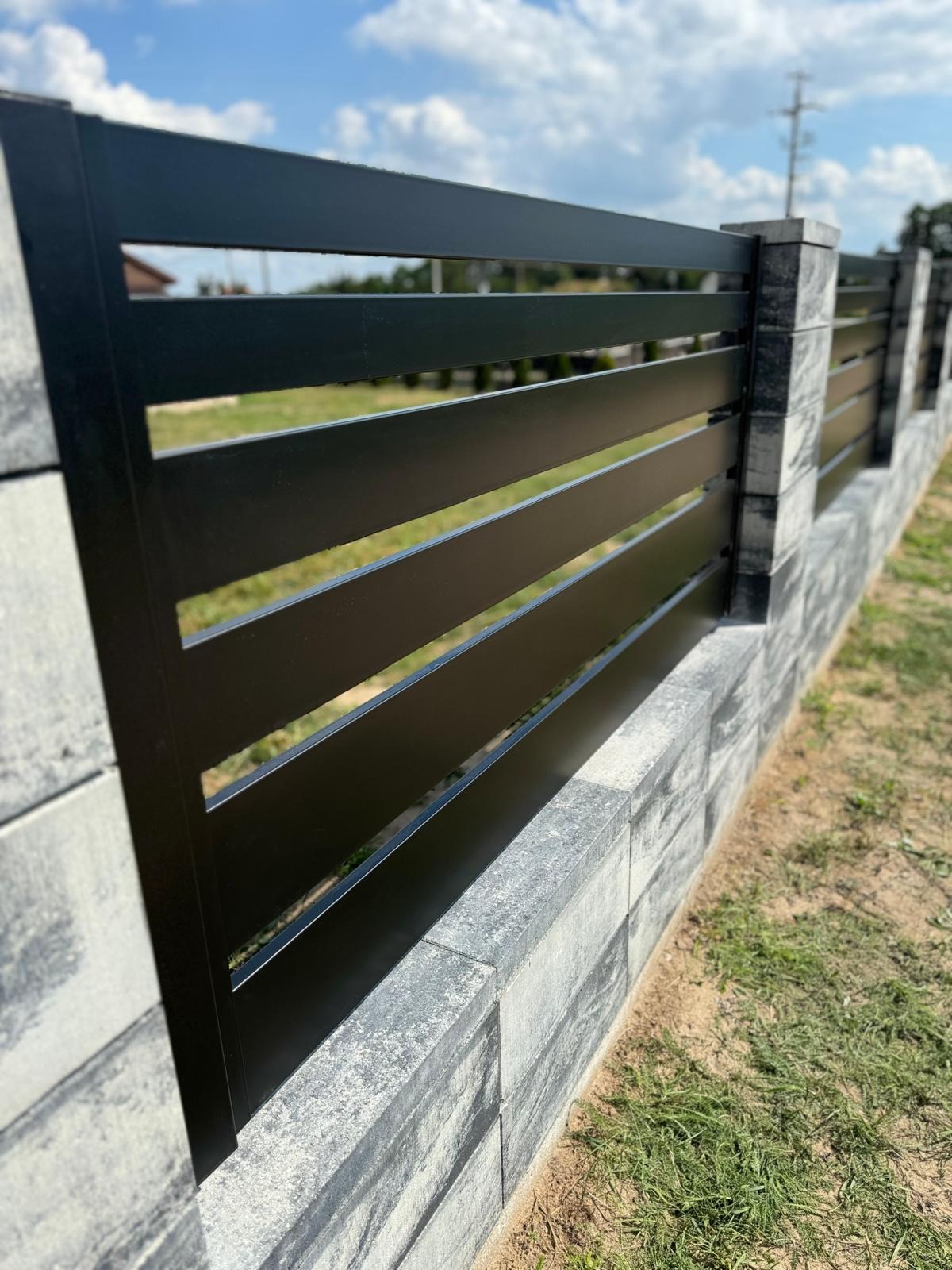 Nowoczesne ogrodzenie panelowe z poziomych, czarnych profili zamontowane na murku z szarych, dekoracyjnych bloczków betonowych, widok z boku w słoneczny dzień.