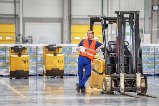 Pracownik w pomarańczowej kamizelce i niebieskich spodniach opiera się o wózek widłowy, trzymając kask na tle magazynu pełnego zapakowanych towarów.