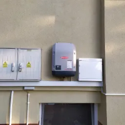 Zewnętrzna instalacja fotowoltaiczna na elewacji budynku: rozdzielnia elektryczna z ostrzeżeniami, inwerter Fronius Symo i dodatkowa skrzynka, wszystko zamontowane na listwie nad oknem.