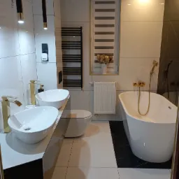 Nowoczesna łazienka z dwoma białymi umywalkami nablatowymi i złotą armaturą, biała wanna wolnostojąca z złotym kranem, czarny grzejnik drabinkowy i czarne akcenty na podłodze i blacie, oświetlenie...