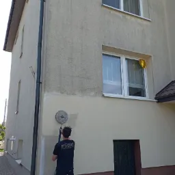 Pracownik w czarnej koszulce z logo firmy czyści elewację budynku za pomocą myjki ciśnieniowej; widoczne różnice w kolorze elewacji przed i po czyszczeniu, okno z niebieską torbą foliową i złoty...
